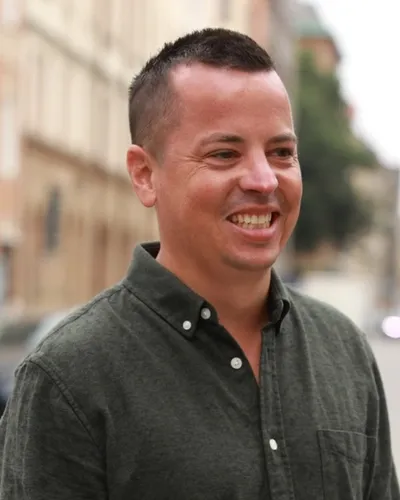 Den leendes coachen och föreläsaren Simon Beyer i mörkgrön skjorta med hus och gata som bakgrund