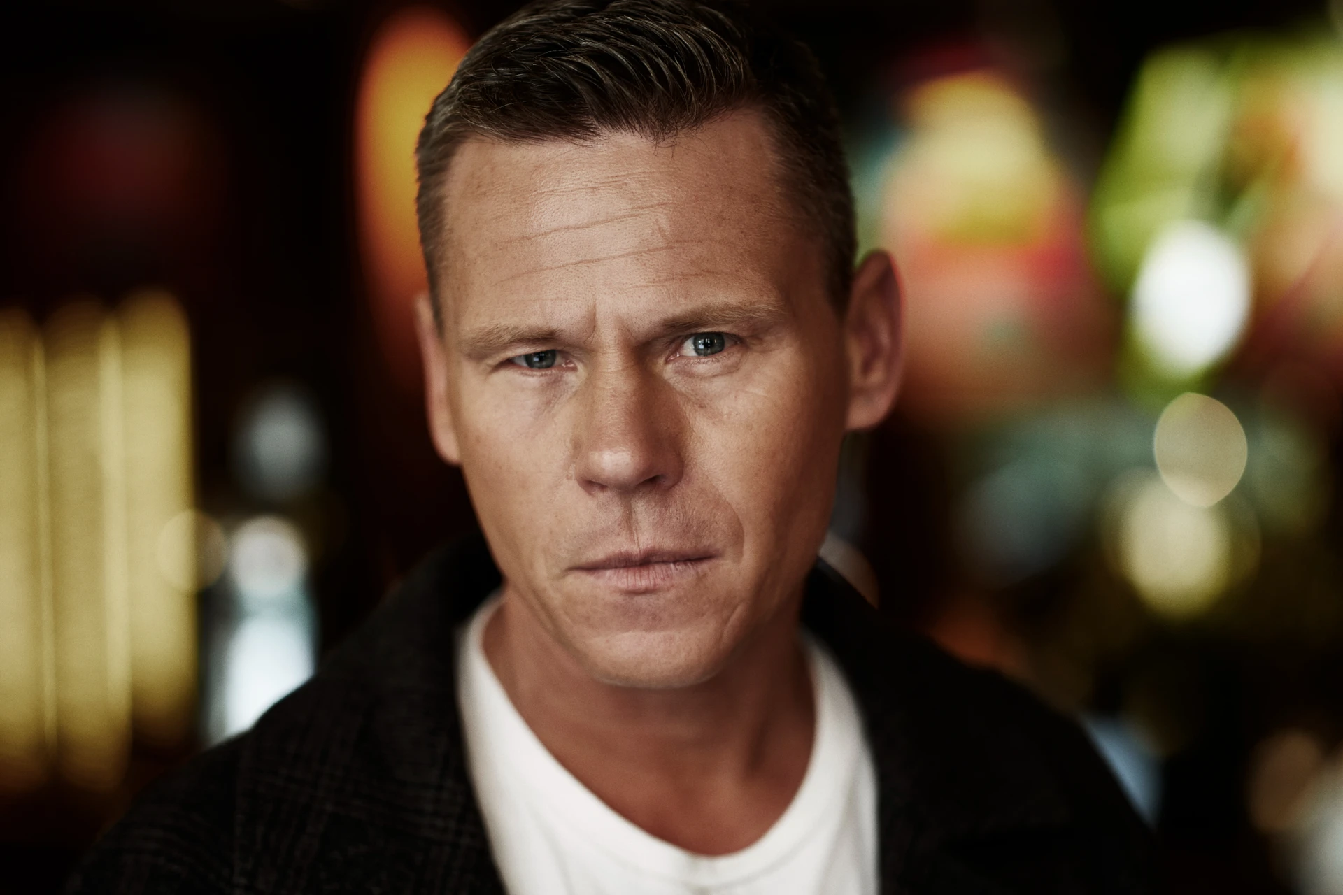 Mons Kallentoft, svensk författare, porträtterad i vit t-shirt och svart öppen kofta, framför suddig innemiljö, för intervju på BYWRTRS