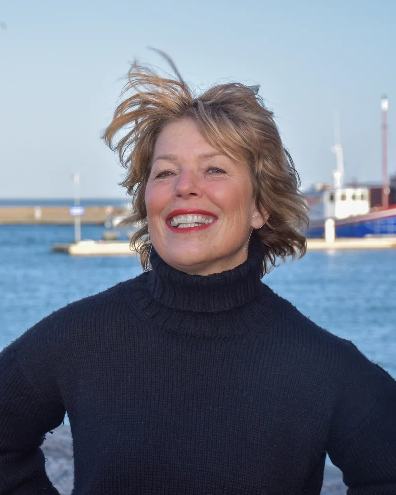 Författaren och kommunikatören Malin Lundskog står framför ett blått hav på ön Knippla, klädd i svart polotröja - med ett stort leende på läpparna.