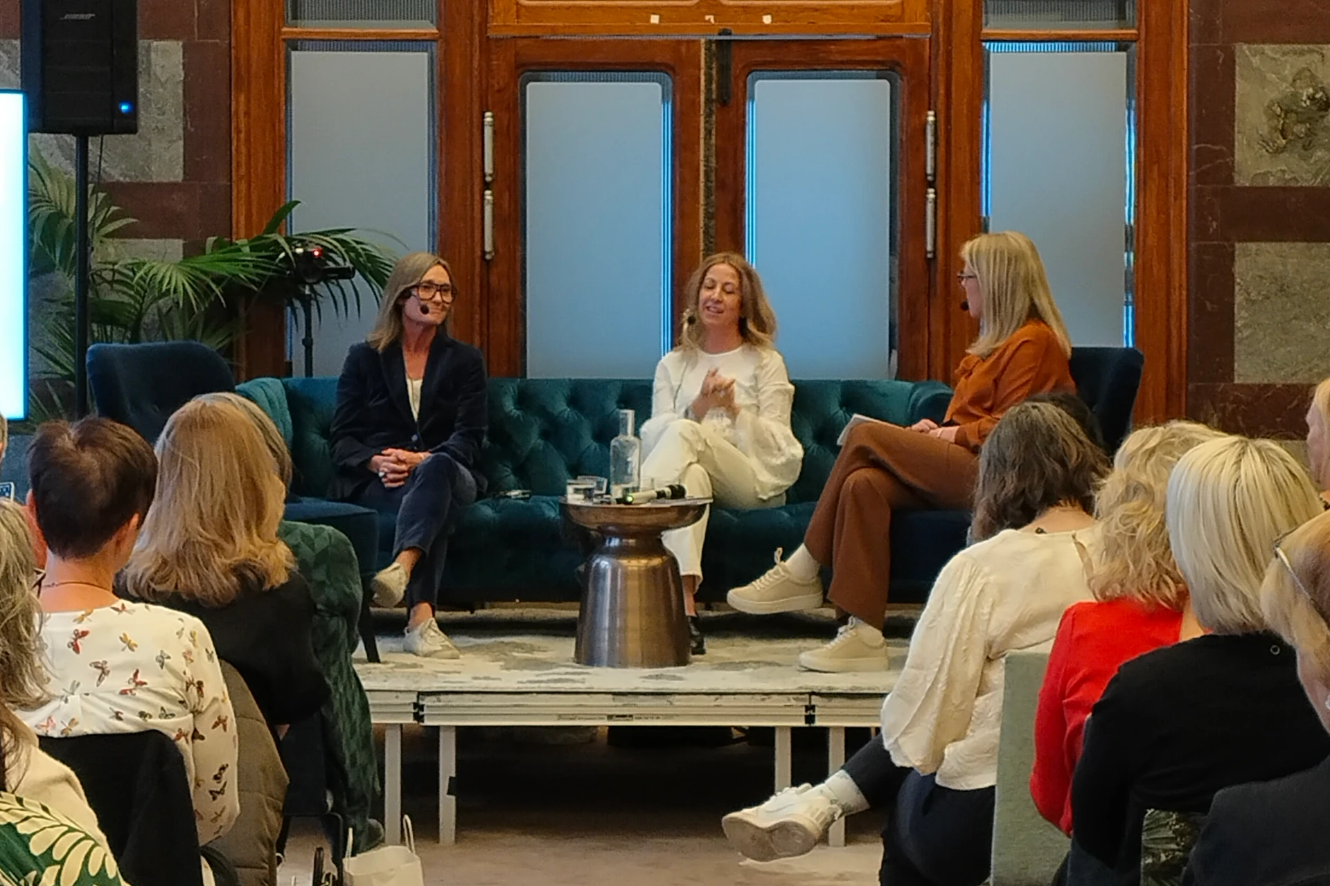 Anna-Lena Hernvall i mörkblå kostym och Alexandra Appelqvist i vit dress från BYWRTRS sitter på en blå sammetssoffa på ett podie under en intervju med Own Your Book. Publik sitter på vardera sida av mittengången, och bakom dem syns stora glaspartier och trädörrar.