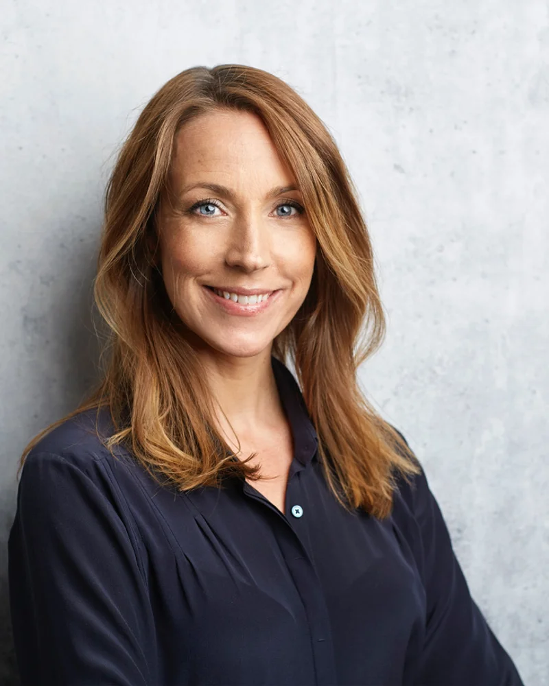 Bilden visar psykologen och författaren Anna Bennich, stående mot en ljusgrå vägg, leende i en blå blus.