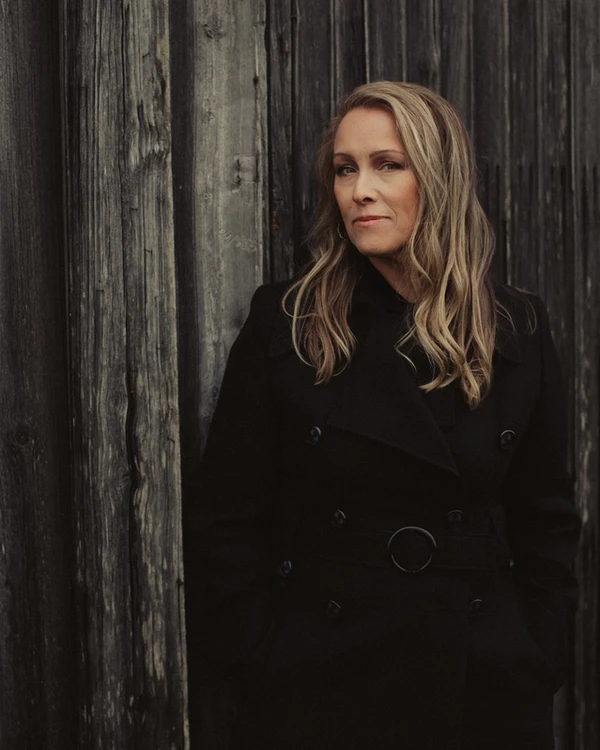 En mörkklädd kvinna med långt hår står framför en vägg av träd.