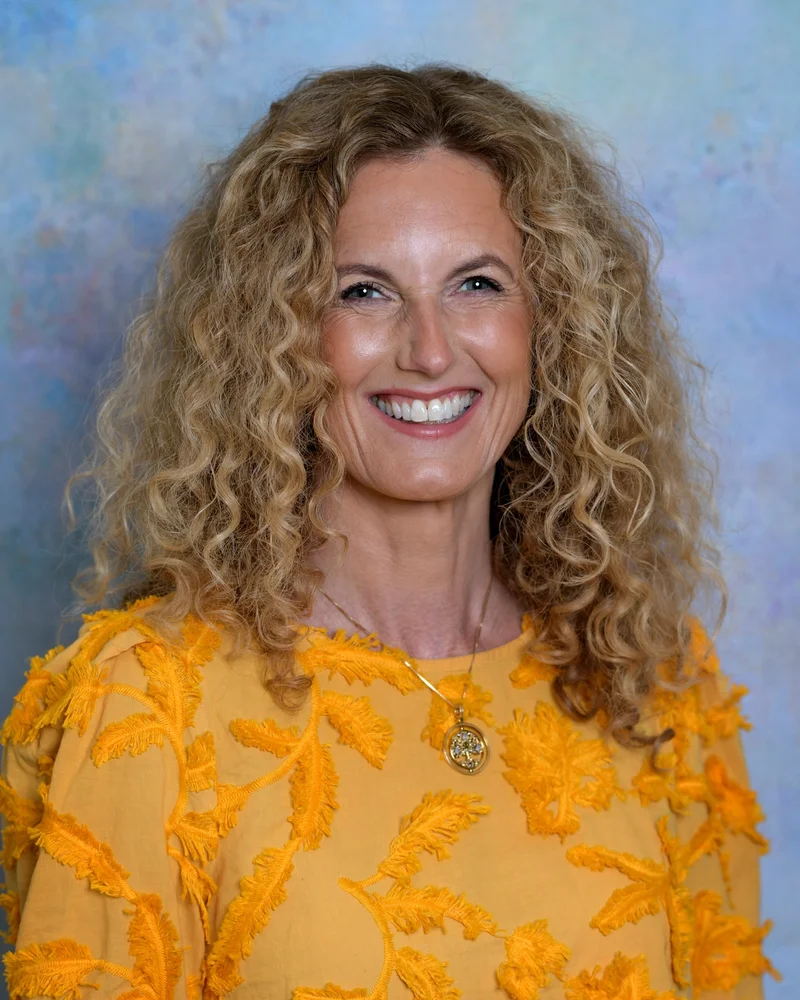 Linda Öhling står klädd i en gul tröja och med ett stort leende på läpparna mot en ljusblå vägg.