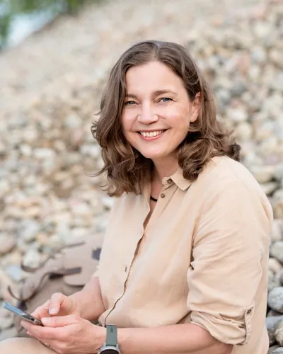 Lena Zetterberg Björk i ljusbeige skjorta sittandes på en sluttning av stenar med natur i bakgrunden.