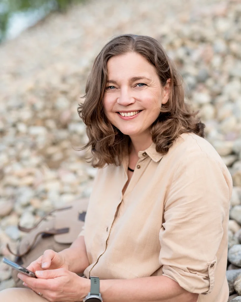 En mörkhårig kvinna står på en stenstrand, klädd i en beige kofta och med ett leende på läpparna.