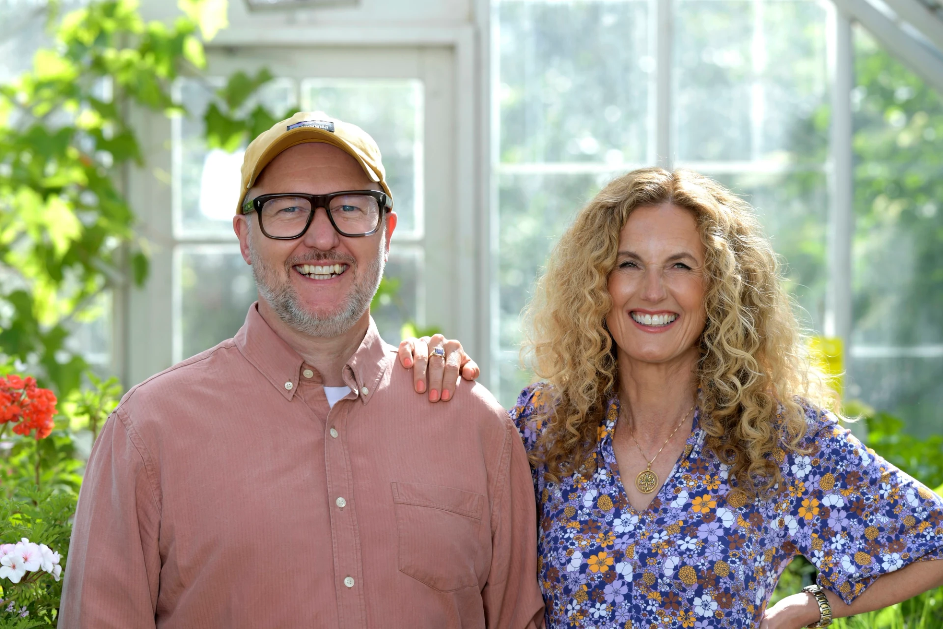 Linda Öhling i blommig blus och Jens Wedin i rostfärgad skjorta står tillsammans och ler mot kameran framför ett växthus.