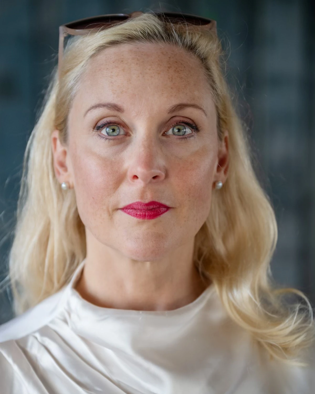 Författaren Lisa Bjurwald ser in i kameran. Hon bär rött läppstift och har solglasögonen i håret.