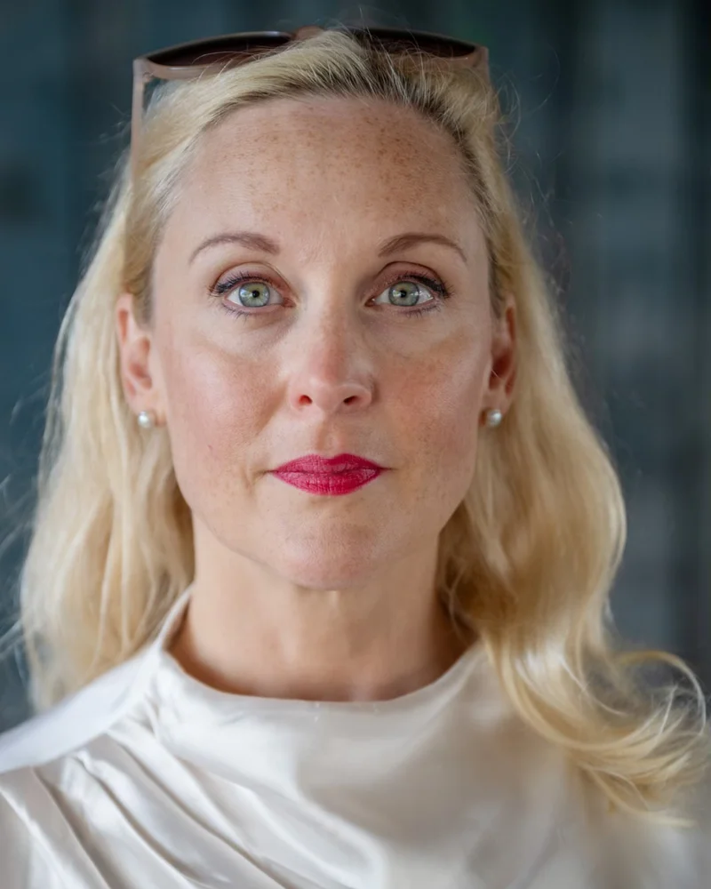 Författaren Lisa Bjurwald ser in i kameran. Hon bär rött läppstift och har solglasögonen i håret.