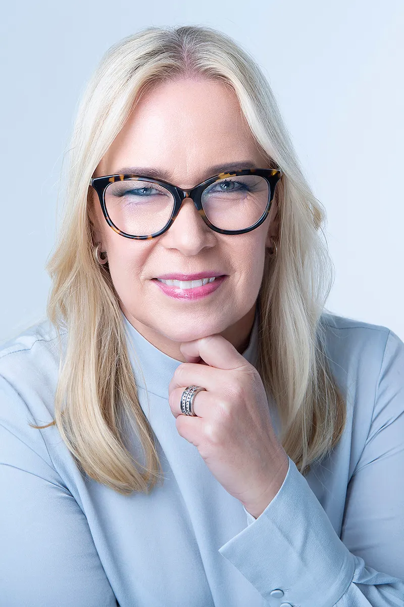 En blond kvinna med svarta glasögon och ljusblå tröja tittar rakt in i kameran. 