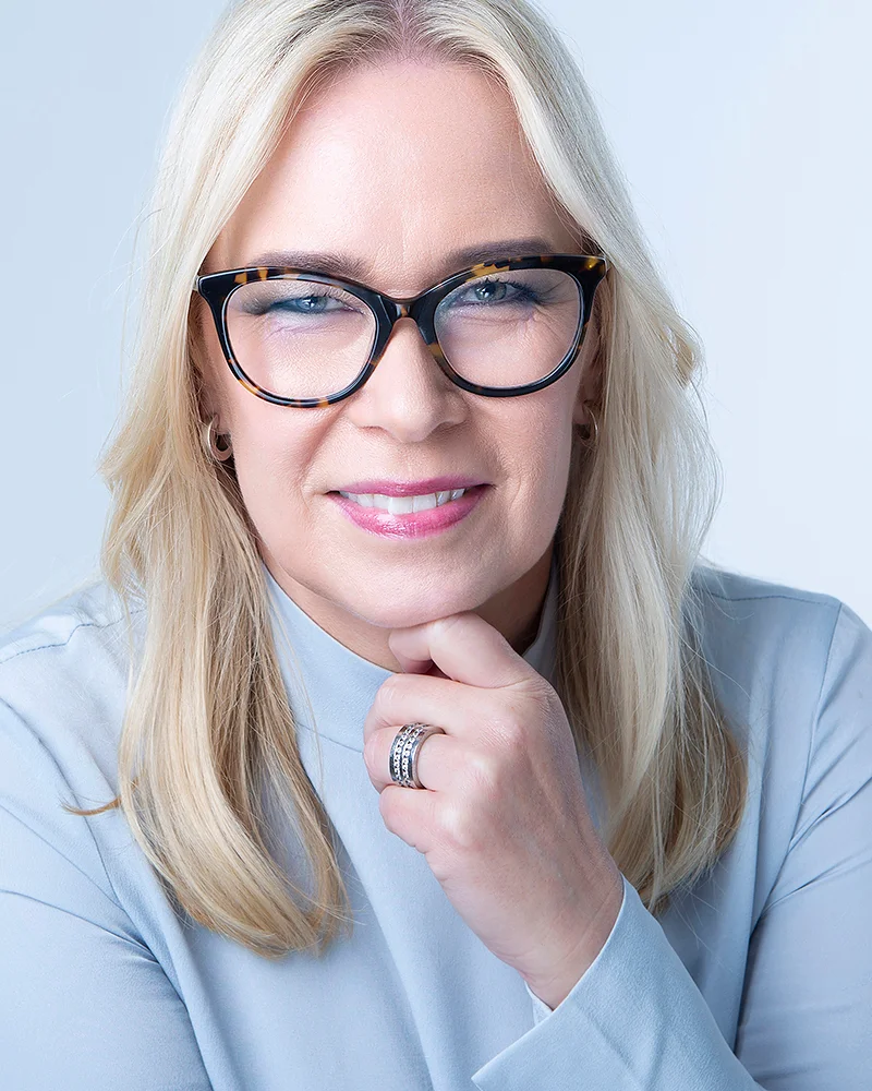 En blond kvinna med svarta glasögon och ljusblå tröja tittar rakt in i kameran.