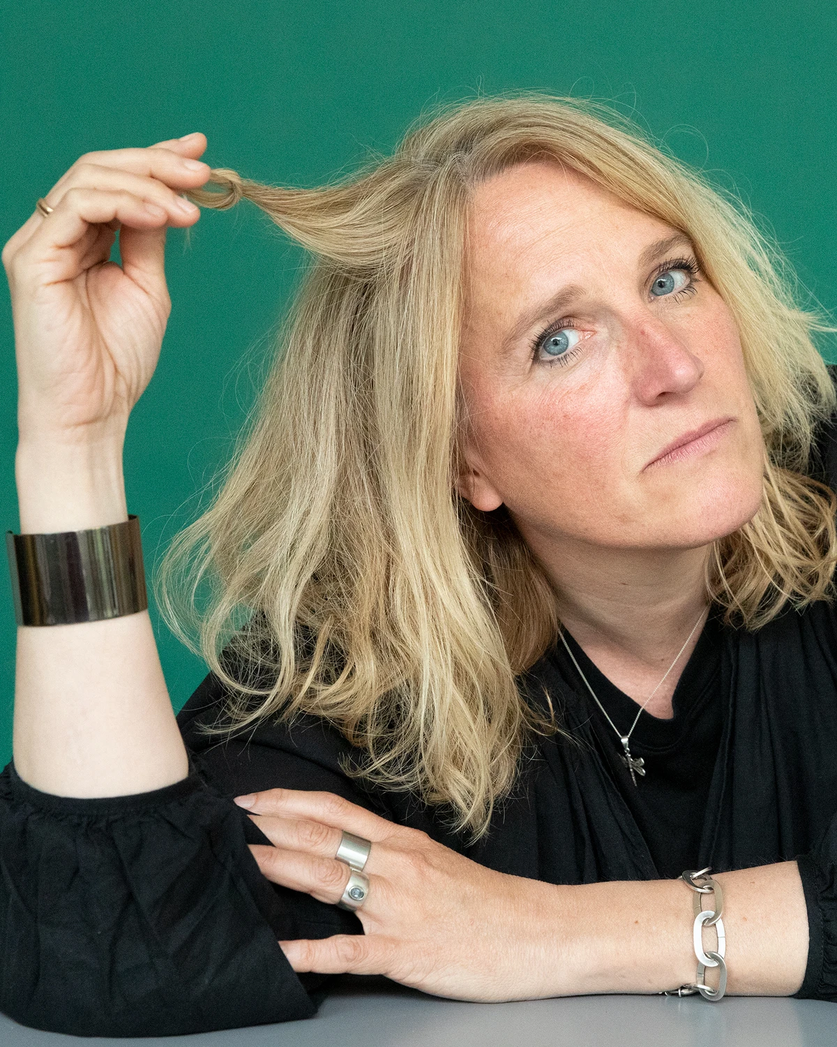 Författaren Frida Spikdotter sitter framför en grön vägg och drar i en hårslinga. Hon har svart blus och stora silversmycken.