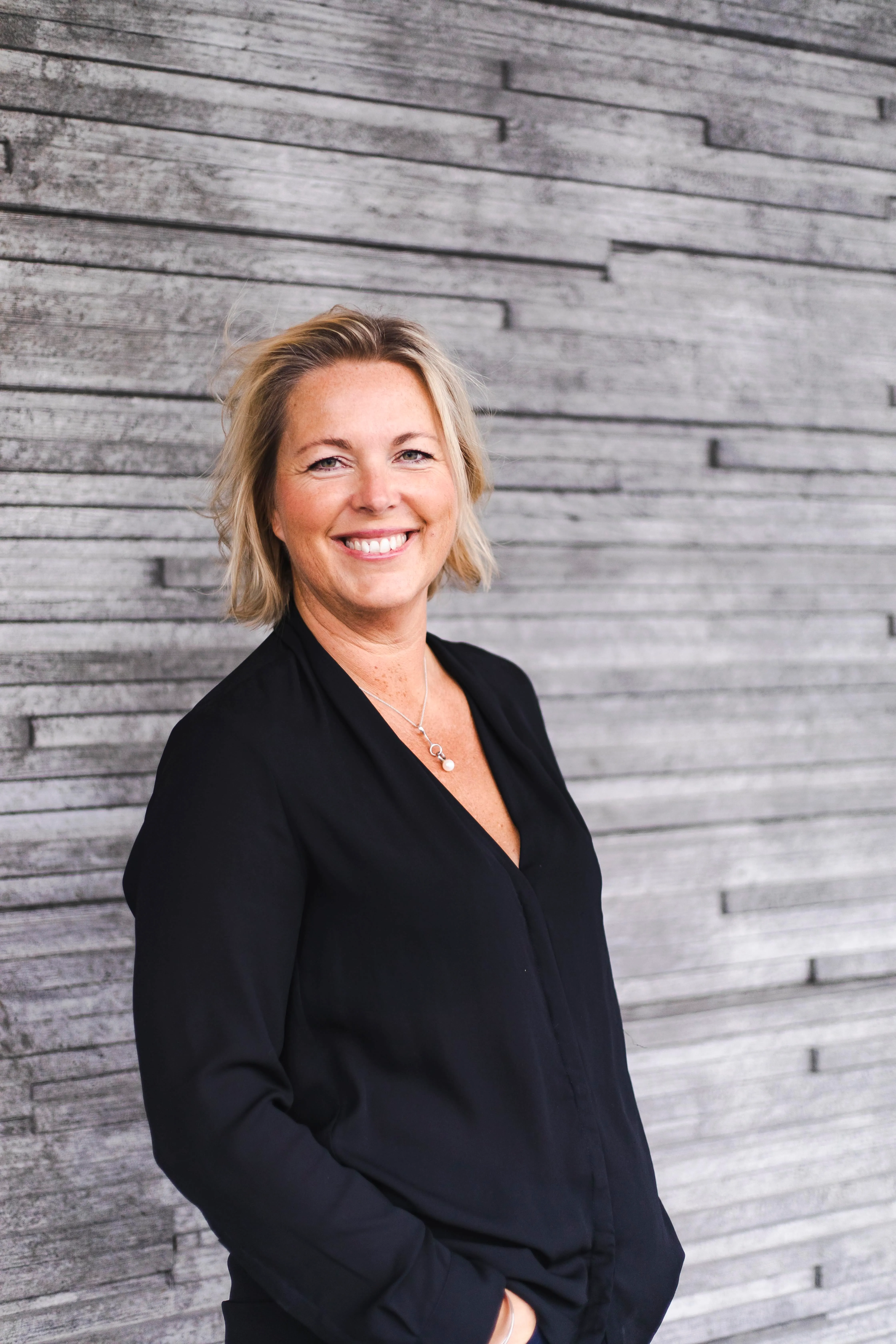 Catarina Kentell står leende i en svart blus framför ett grått plank. 