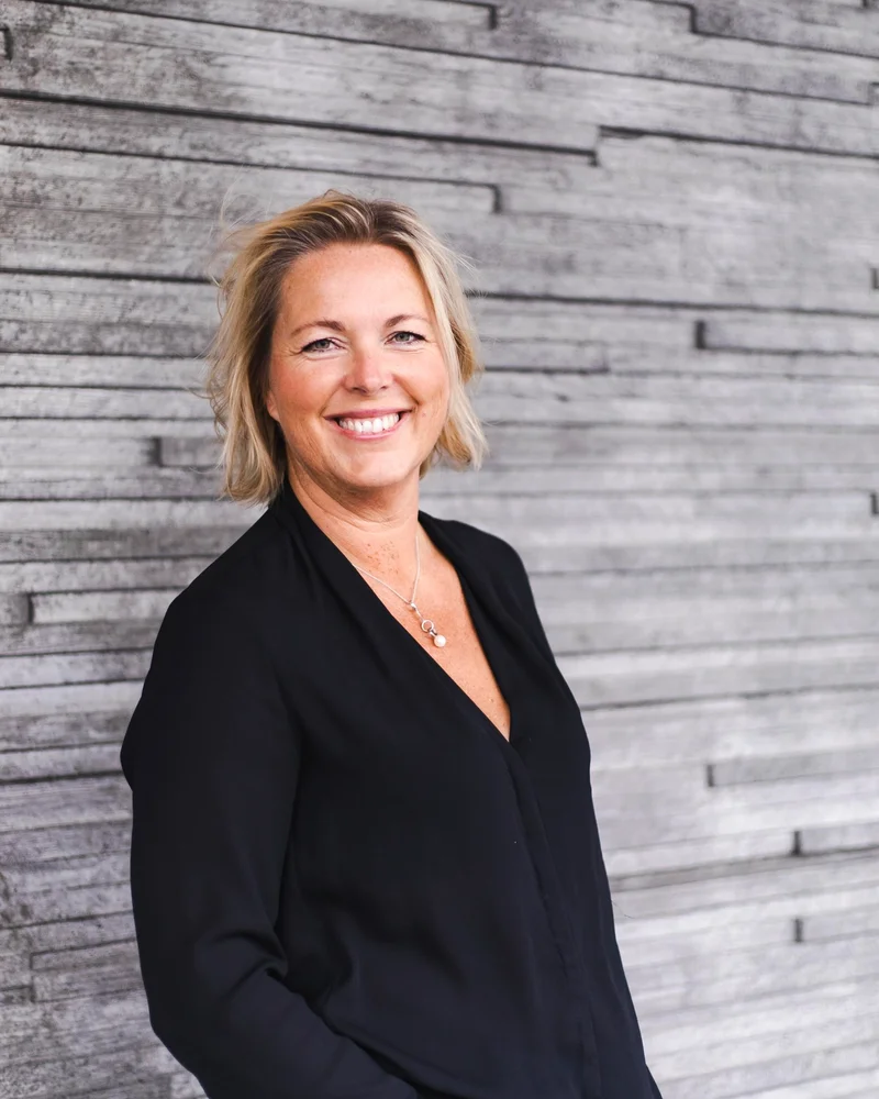 Catarina Kentell står leende i en svart blus framför ett grått plank.