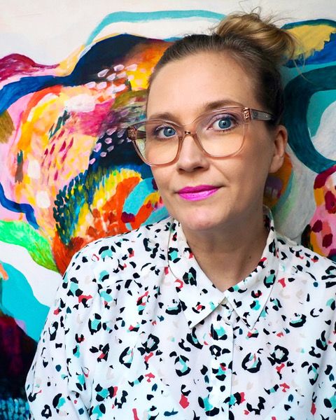 Författaren Jenny Theolin bär glasögon, cerist läppstift och en prickig skjorta. Bakom henne ett färgsprakande konstverk.