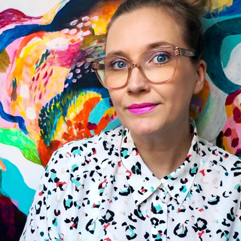 Jenny Theolin bär läppstift i cerise, glasögon och en prickig blus. Hon har håret i en knut och står framför en abstrakt målning