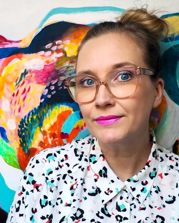 Författaren Jenny Theolin bär glasögon, cerist läppstift och en prickig skjorta. Bakom henne ett färgsprakande konstverk.