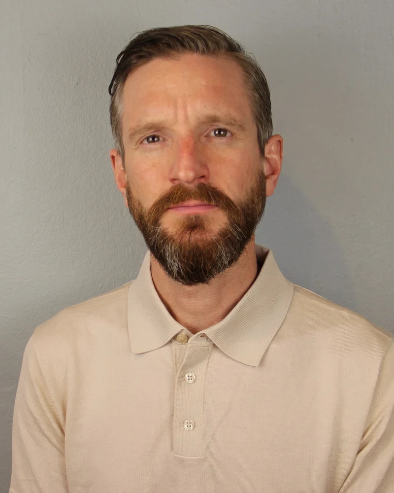 En man i mörkt hår och skägg, klädd i en vit tenniströja, tittar in i kameran.