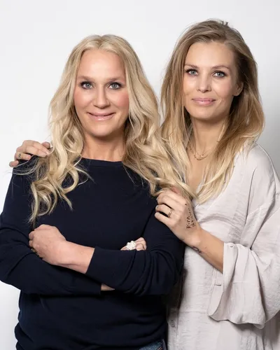 Stjärnduon Ann Söderlund och Fi Lindfors står tätt tillammans och tittar leende in i kameran.