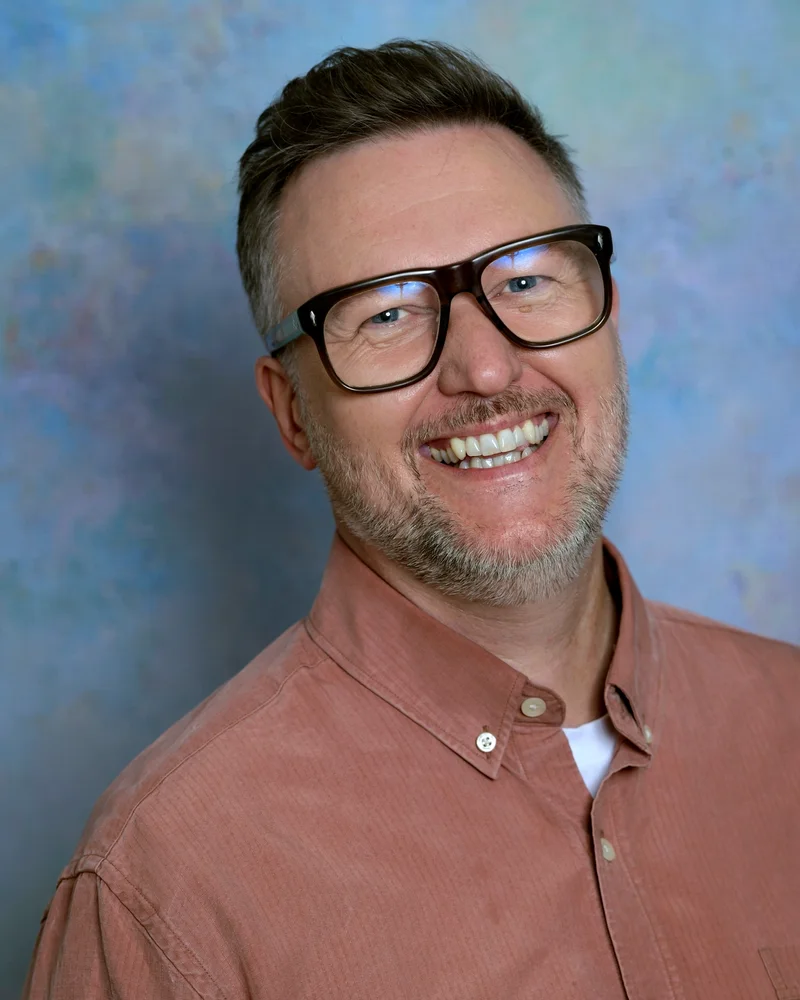 Jens Wedin står mot en ljusblå vägg med ett stort leende på läpparna, glasögon och en mörkrosa skjorta.