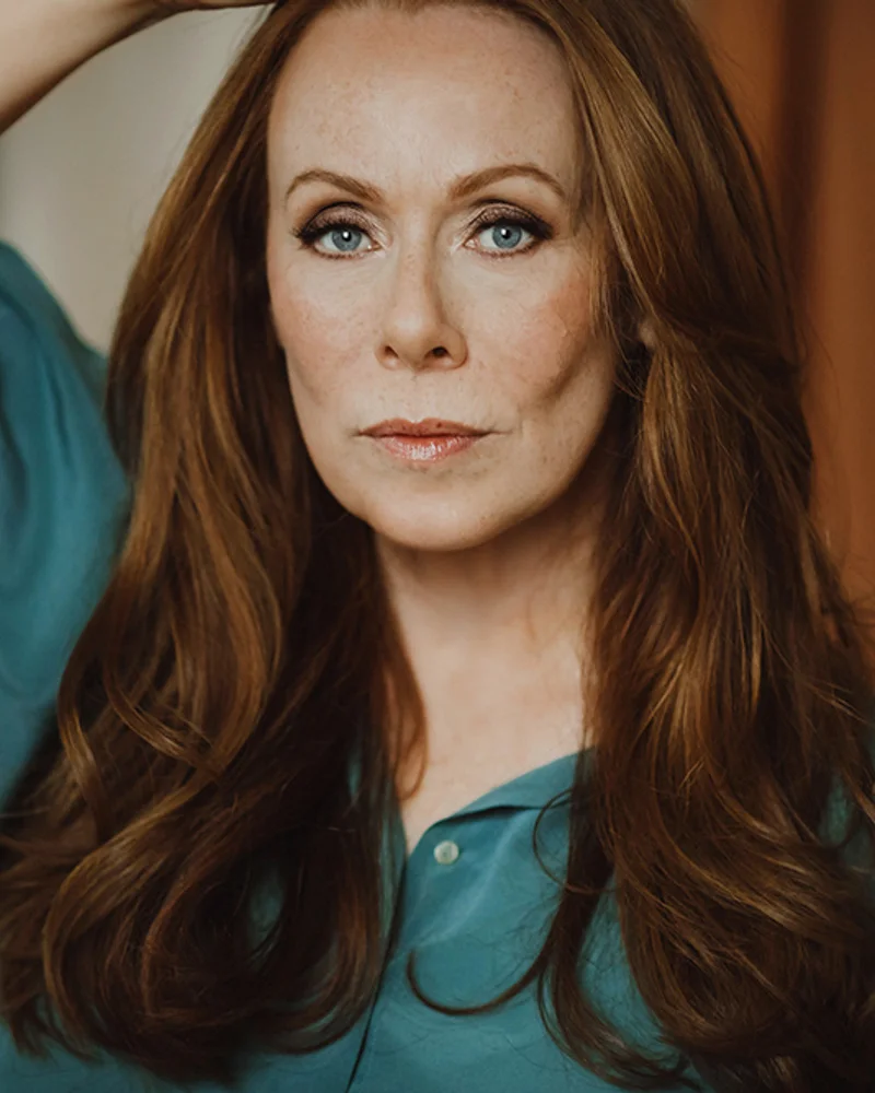 Skådespelare & författare Rachel Mohlin i turkos blus och poserandes med ena armen upp vid huvudet