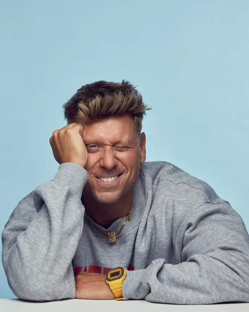 Artisten och författaren Johnny Johansson sitter lutad över en bordsskiva och vilar huvudet mot sin hand. Klädd i grå sweatshirt och med en klurig grimas i ansiktet.