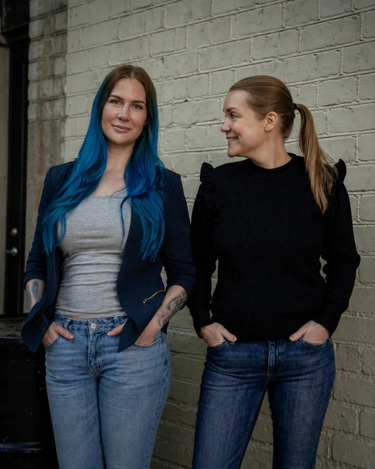 Författarna och föreläsarna Mariah Eriksson och Jenny Hamrén står bredvid varandra framför en tegelvägg.