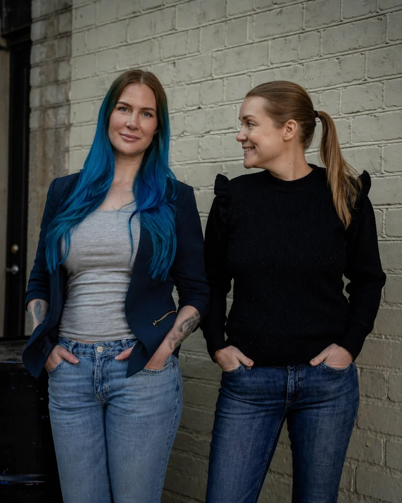 Författarna och föreläsarna Mariah Eriksson och Jenny Hamrén står bredvid varandra framför en tegelvägg.