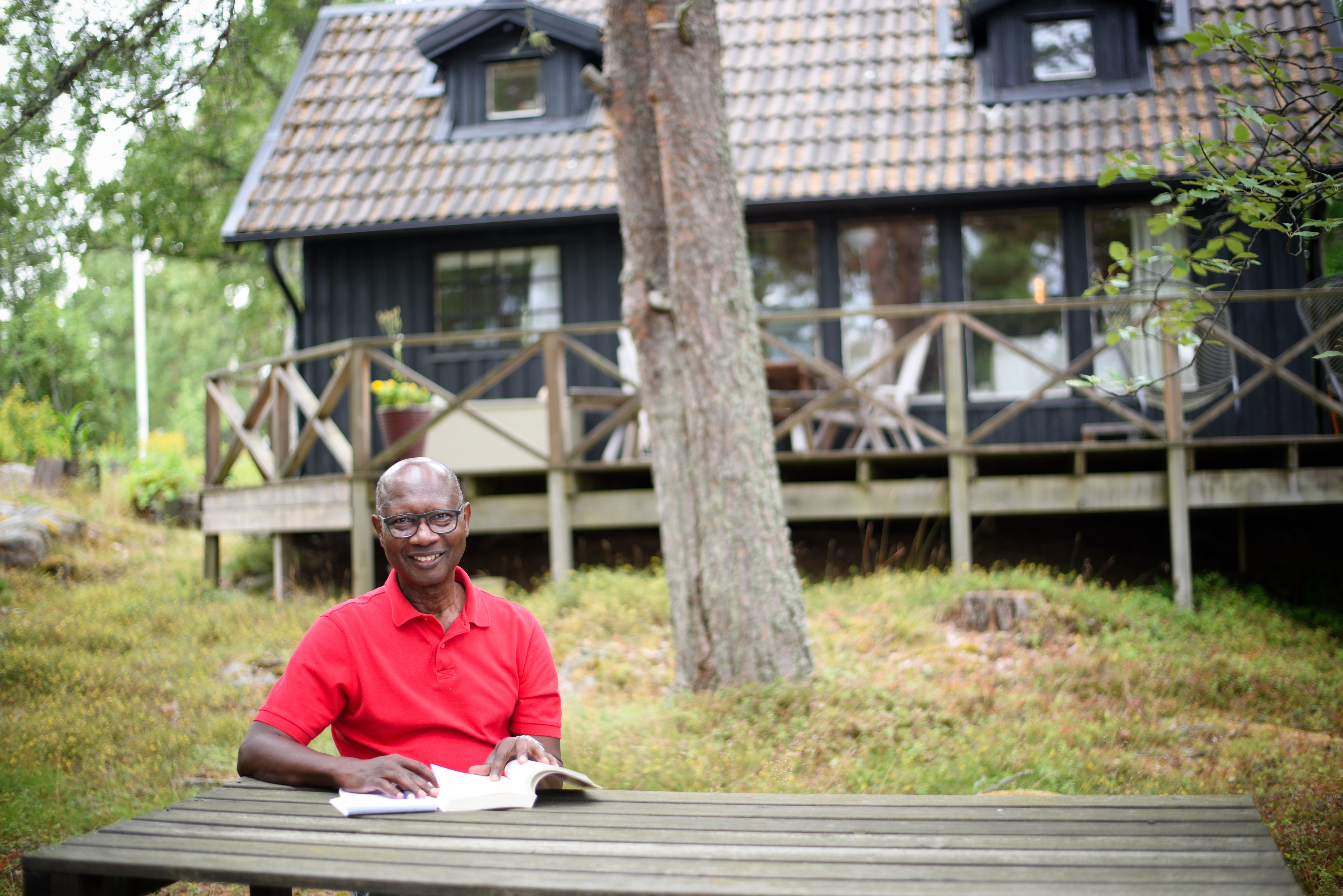 Författaren Amadou Jallow sitter i röd tröja framför sitt skärgårdshus på Gräsö