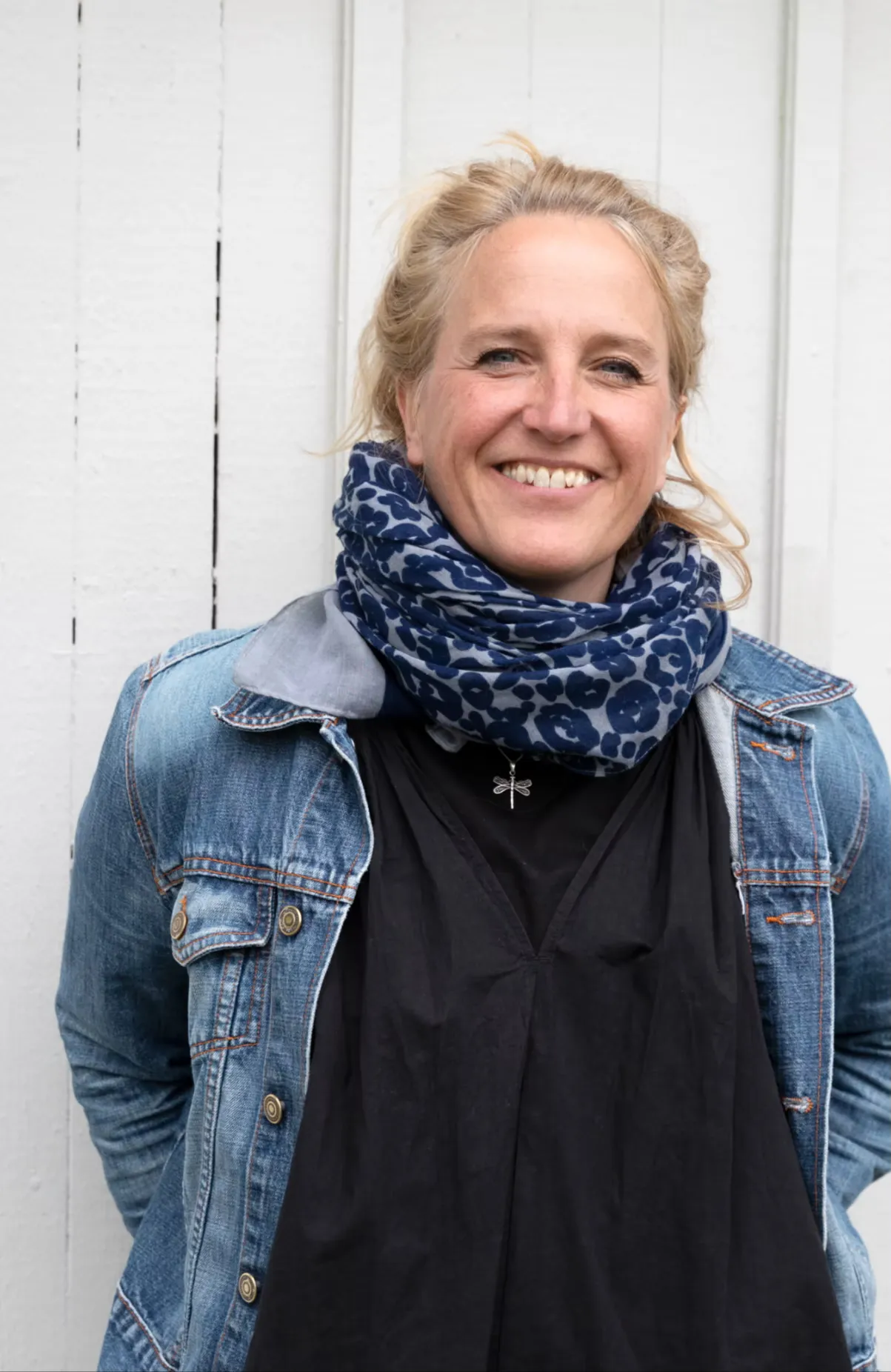En blond kvinna klädd i svart tröja, jeansjacka och en blå schal står vid en vit vägg med ett stort leende på läpparna. 