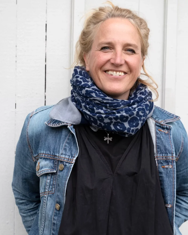 En blond kvinna klädd i svart tröja, jeansjacka och en blå schal står vid en vit vägg med ett stort leende på läpparna.