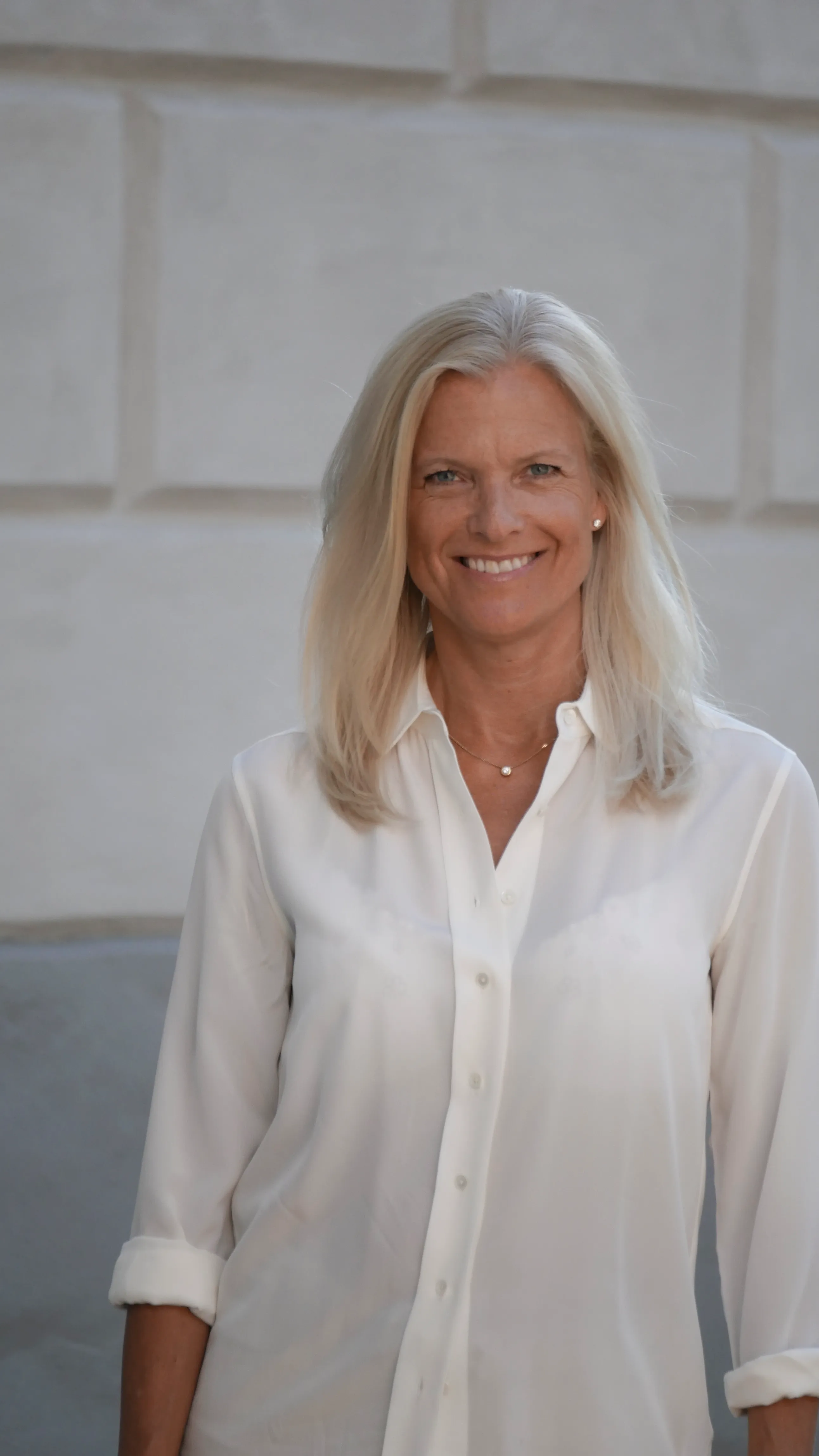 En blond kvinna klädd i vit blus står framför en vägg med ett leende. 