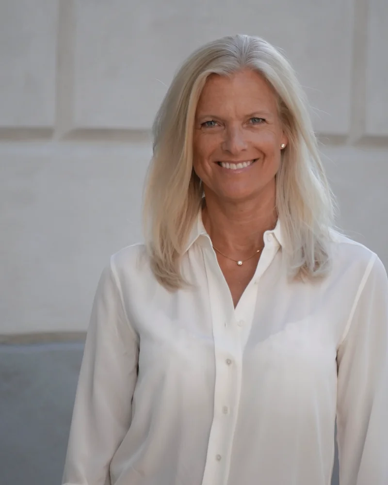 En blond kvinna klädd i vit blus står framför en vägg med ett leende.