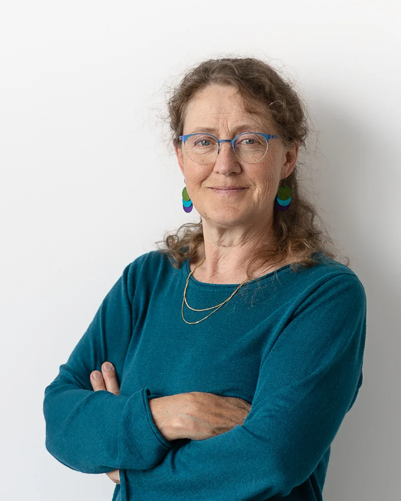 En kvinna med långt hår, glasögon och klädd i turkosblå tröja står framför en ljus vägg.