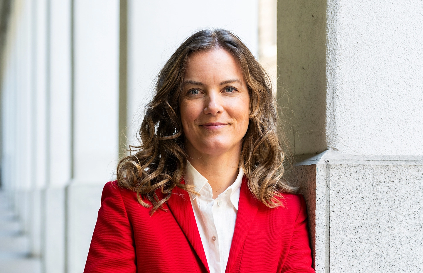 Författaren Nina Modig står lutad mot en grå och vit marmorpelare i en utomhuspelargång. Hon ler mot kameran och bär en vit skjorta och en röd kavaj.