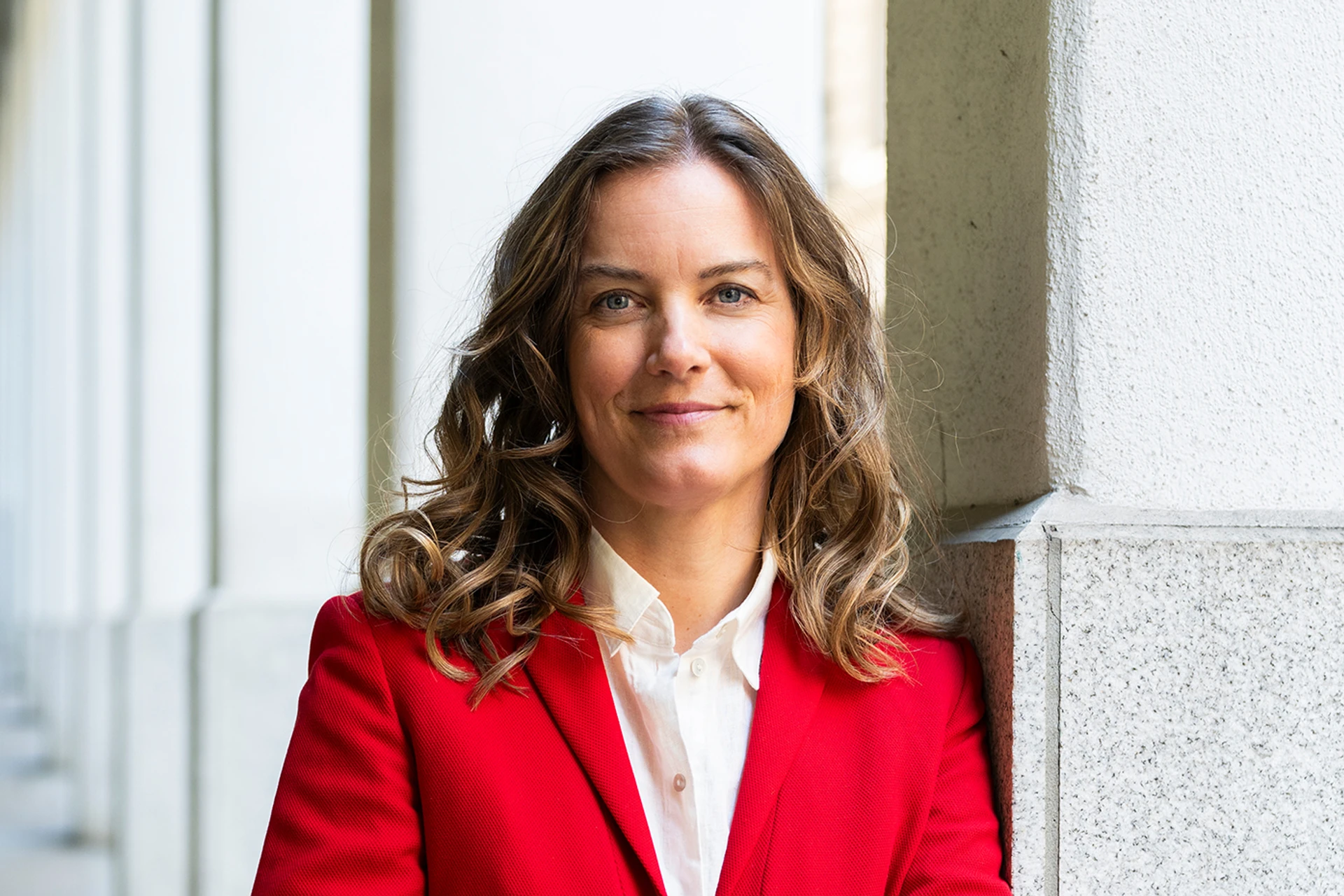 Författaren Nina Modig står lutad mot en grå och vit marmorpelare i en utomhuspelargång. Hon ler mot kameran och bär en vit skjorta och en röd kavaj.