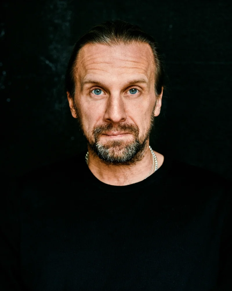 En mörkhårig man med skägg blickar rakt in i kameran. Klädd i svart tröja.