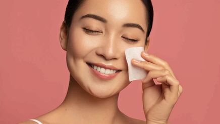 A smiling Asian woman using a cotton pad to apply skincare.