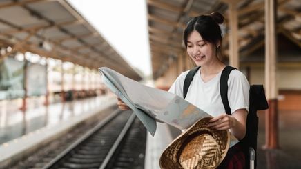 Asian woman is at a railway station looking at a map with a hat in one hand.
