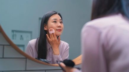 Asian woman applying foundation with a sponge in front of a mirror.