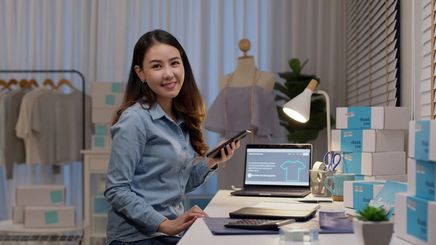 Smiling Asian woman working and holding a tablet with a visible laptop and clothing rack.