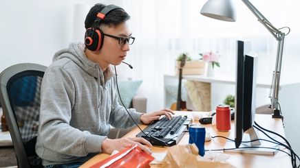 A young Asian man playing games on his computer using a complete headset