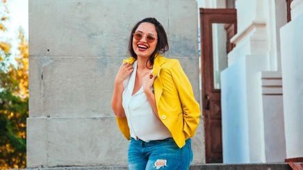 Smiling woman wearing ripped denim pants, sunglasses, and yellow blazer.
