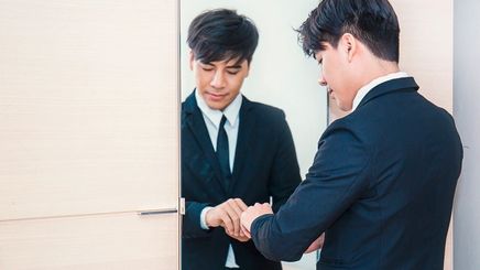 Asian man in a suit, fixing cuffs in front of mirror.