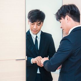 Asian man in a suit, fixing cuffs in front of mirror.