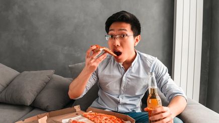 Asian man with glasses eating pizza and drinking beer on couch.