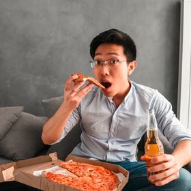 Asian man with glasses eating pizza and drinking beer on couch.