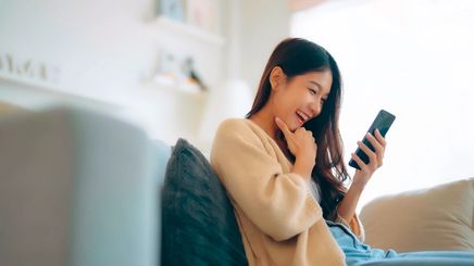Asian woman laughing while looking at phone on couch.