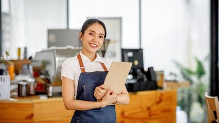 Asian businesswoman wearing an apron and holding a clipboard at a café.