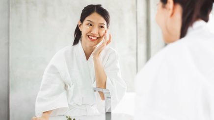 Asian woman in a robe washing face in front of a mirror