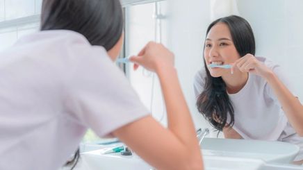 Young Asian woman brushing her teeth in front of a mirror.